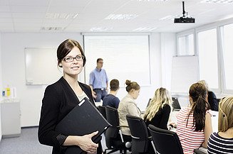 Frau steht mit Tablet in der Hand im Vordergrund, im Hintergrund sitzen Kolleg:innen in einem Konferenzraum.                  