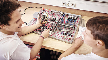 Foto zwei Arbeiter vor Schaltungsplatte, einer mit Messgerät in der Hand