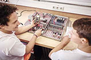 Foto zwei Arbeiter vor Schaltungsplatte, einer mit Messgerät in der Hand