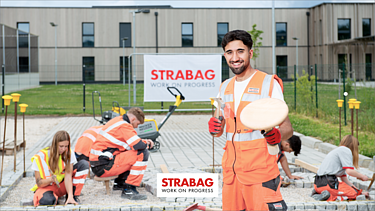 Foto Pflasterer auf Parkplatz, im Hintergrund verlegen Kolleginnen und Kollegen Steine